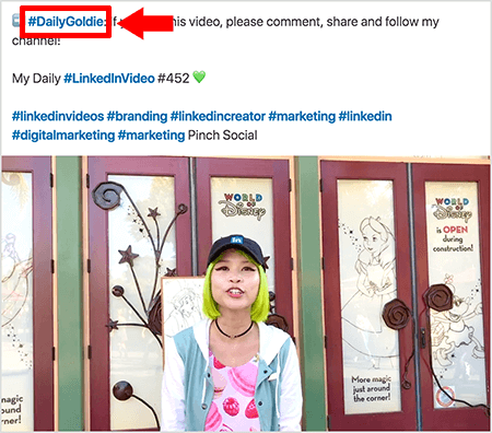 To je posnetek zaslona, ​​ki ponazarja, kako Goldie Chan uporablja hashtago v besedilu svojih videoposnetkov v LinkedInu. Rdeči oblački kažejo na hashtag #DailyGoldie v besedilu, ki je edinstven za njene video objave in ji pomaga slediti skupnim rabam. Objava vključuje tudi druge ustrezne hashtagove, ki ljudem pomagajo najti njen video, med drugim tudi #LinkedInVideo. Na video podobi Goldie stoji pred nekaterimi vrati na razstavi World of Disney. Je Azijka z zelenimi lasmi. Oblečena je v črno kapo LinkedIn, črno ogrlico z ogrlico, roza majico s tiskanim makaronom in modro-belo jakno.